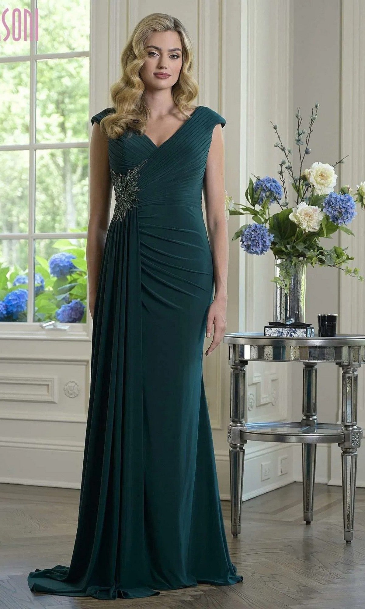 A woman wears the Marsoni M343 Long Formal Dress in deep green, featuring a ruched fitted bodice and decorative brooch, standing in an elegant room with large windows and a floral table arrangement.